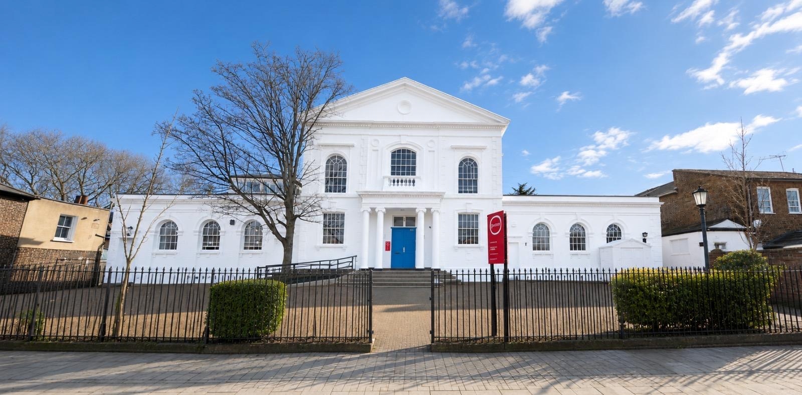 Exterior CGI of Partou Norwood Day Nursery & Pre-School, a new nursery opening in West Norwood, London in October 2026. The image shows a grand white nursery building with tall arched windows, a bright blue front door, red Partou signage, black railings, green landscaping and a sunny blue sky, creating a welcoming and contemporary first impression for families.