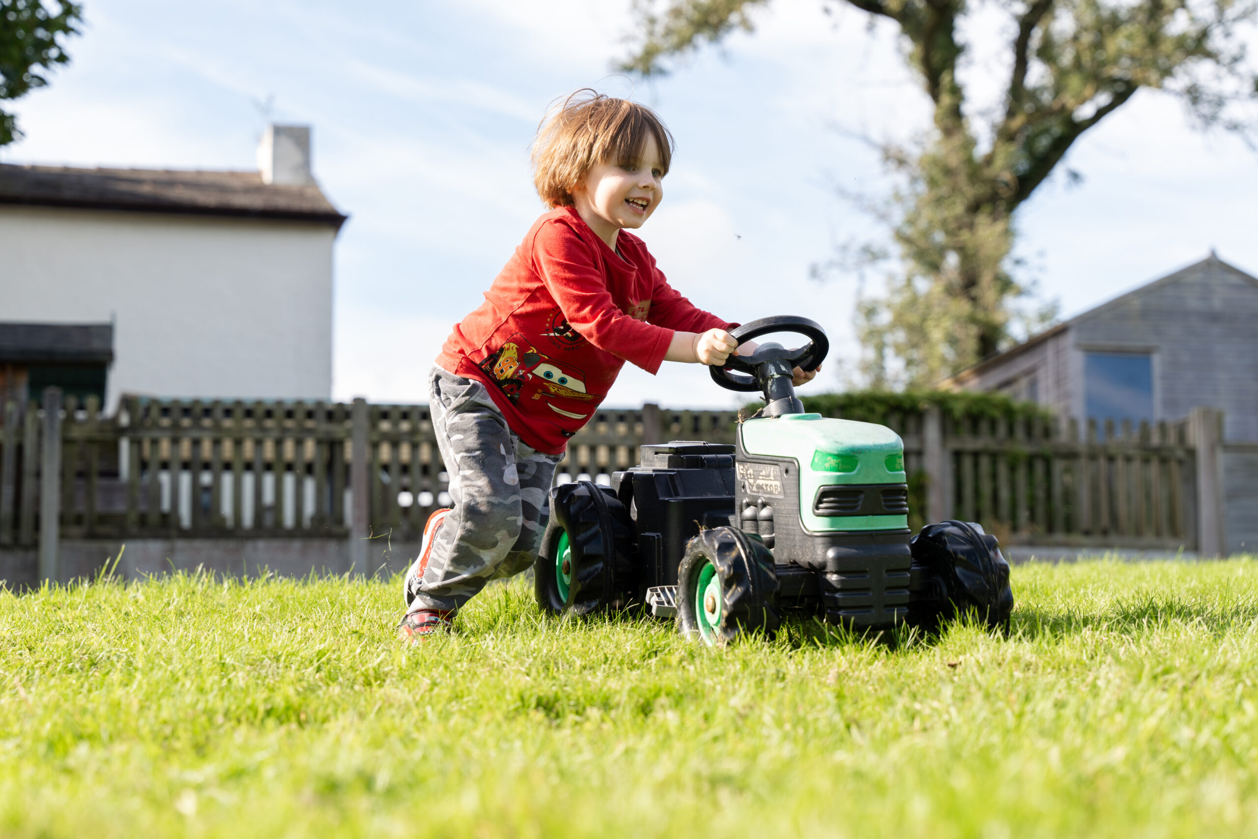 Smiling child with red t-shirt and camoflage trousers pushing a small tractor toy