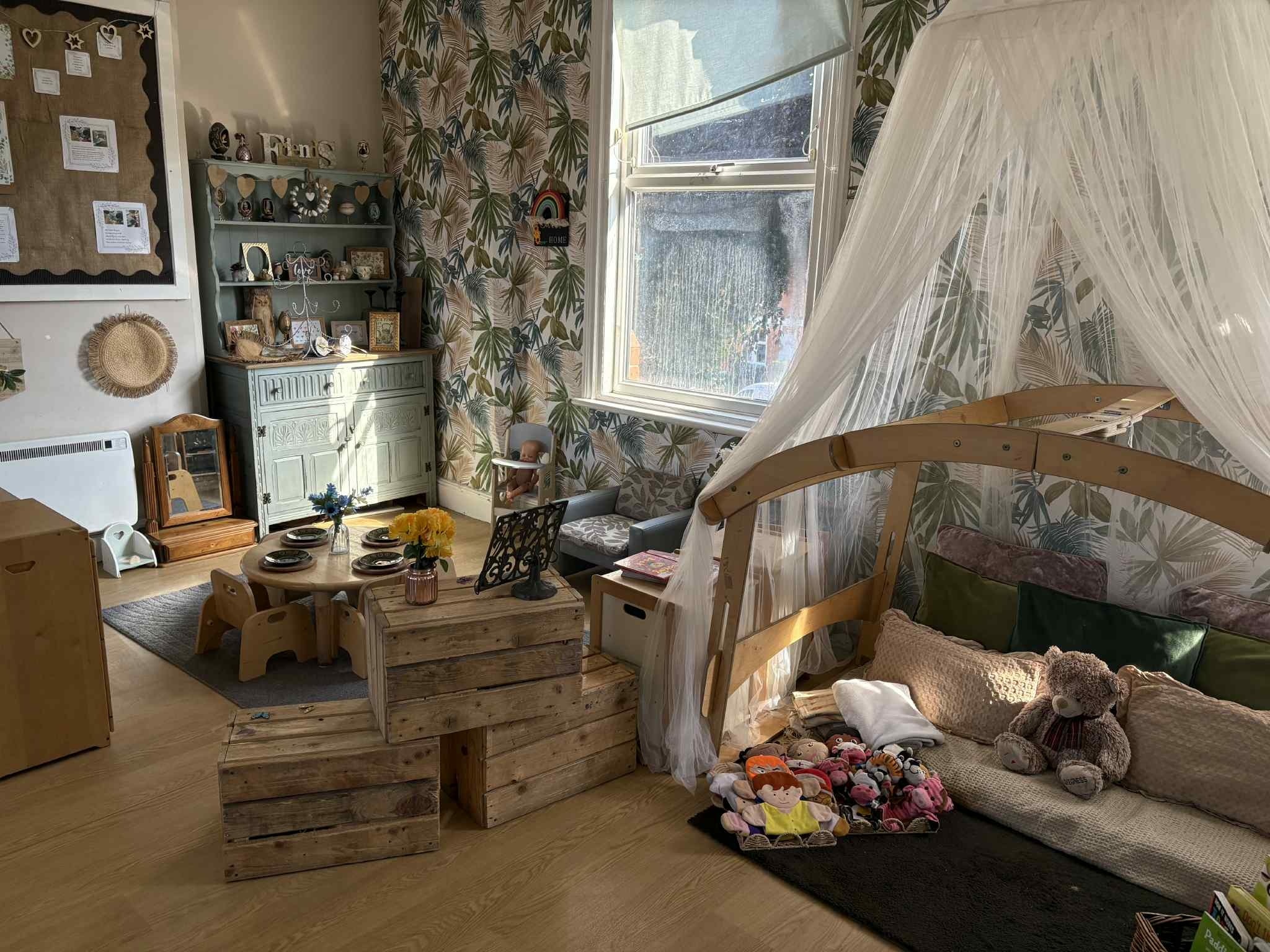 Image of green and print tropical leaf print wall paper with wooden toys and cosy cushioned reading area covered with a white canopy at Partou Arnold Day Nursery