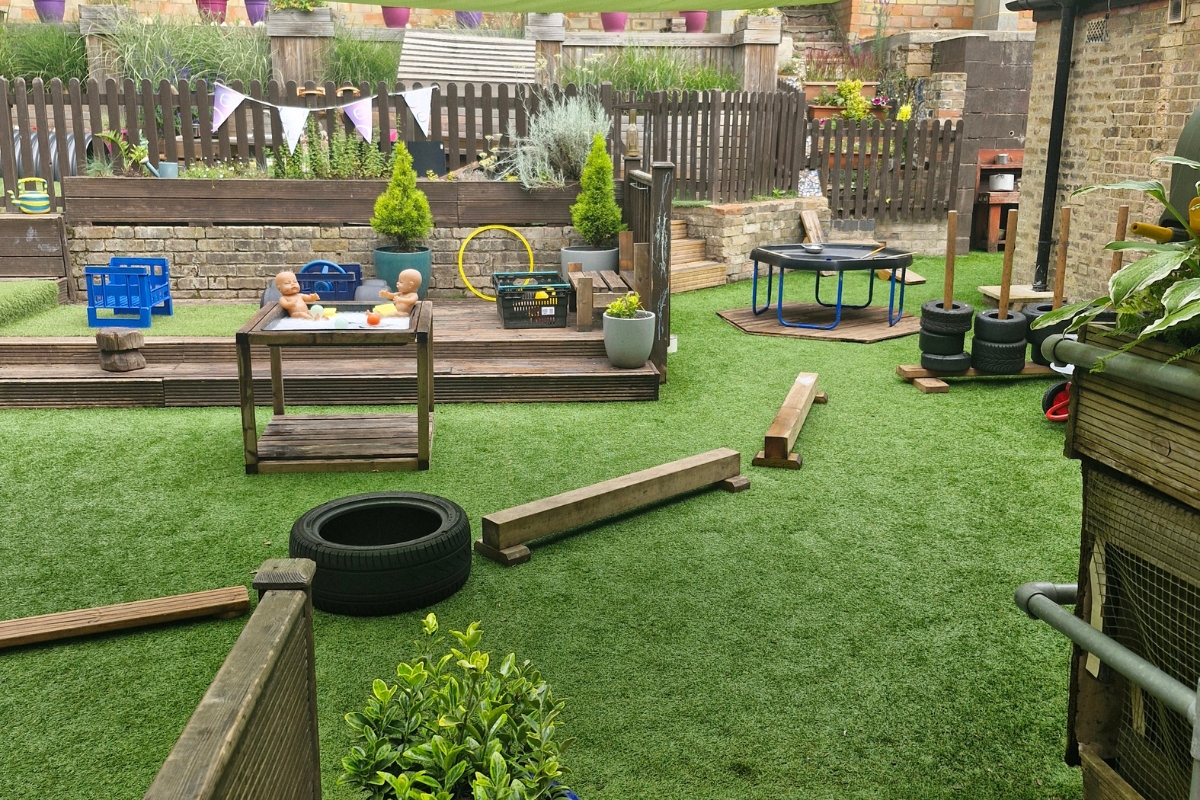 Outdoor play area at Partou Sycamore House Day Nursery with artificial grass, wooden balance beams, a tyre and small trampoline, a water‑play table with dolls and toy fruit, crates, hoops and potted plants within a fenced, bunting‑decorated space