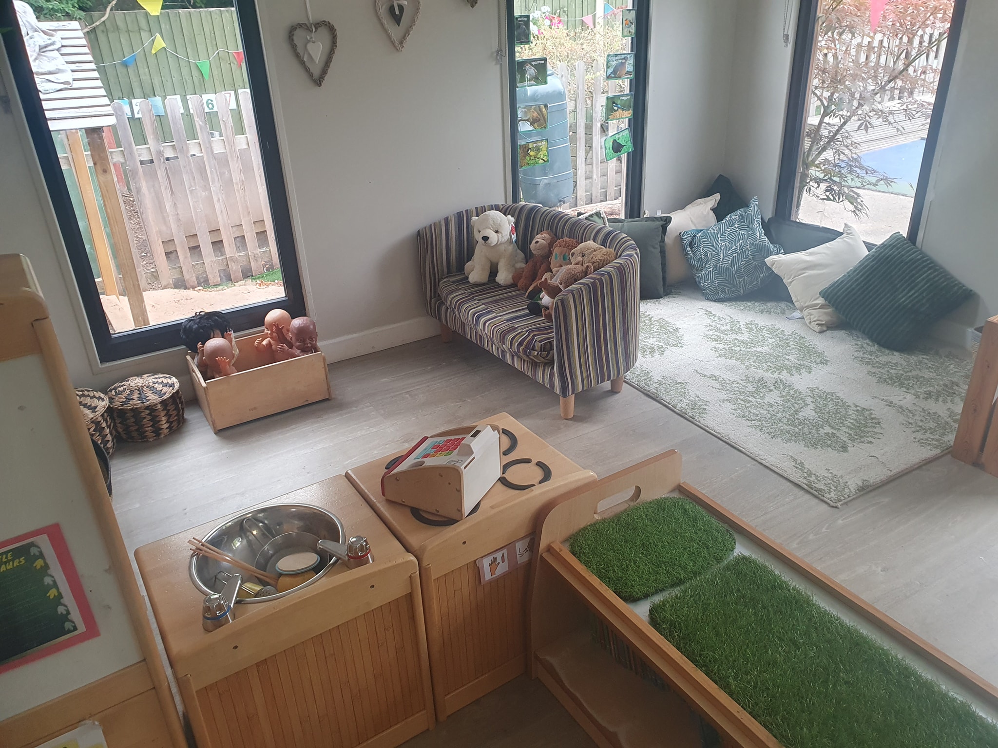 Wooden sink and kitchen hob toys near to a sofa covered with teddies and a rug with cushions at Partou Scotts Wood day nursery in West Bridgford