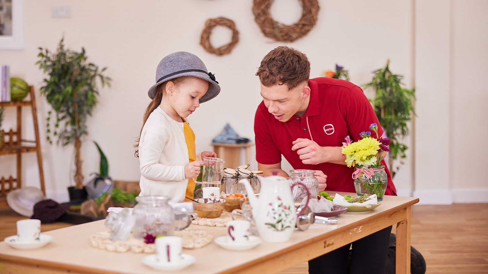 Young girl wearing a grey hat, whilst pouring liquid into jars. Nursery worker with a red Partou polo shirt sat at the table with the young girl.