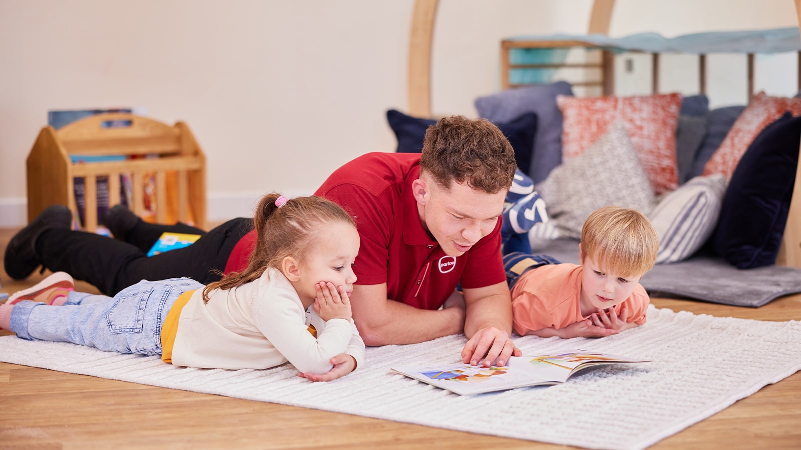 Male nursery worker in a red Partou branded polo shirt, lying down reading a book with two children