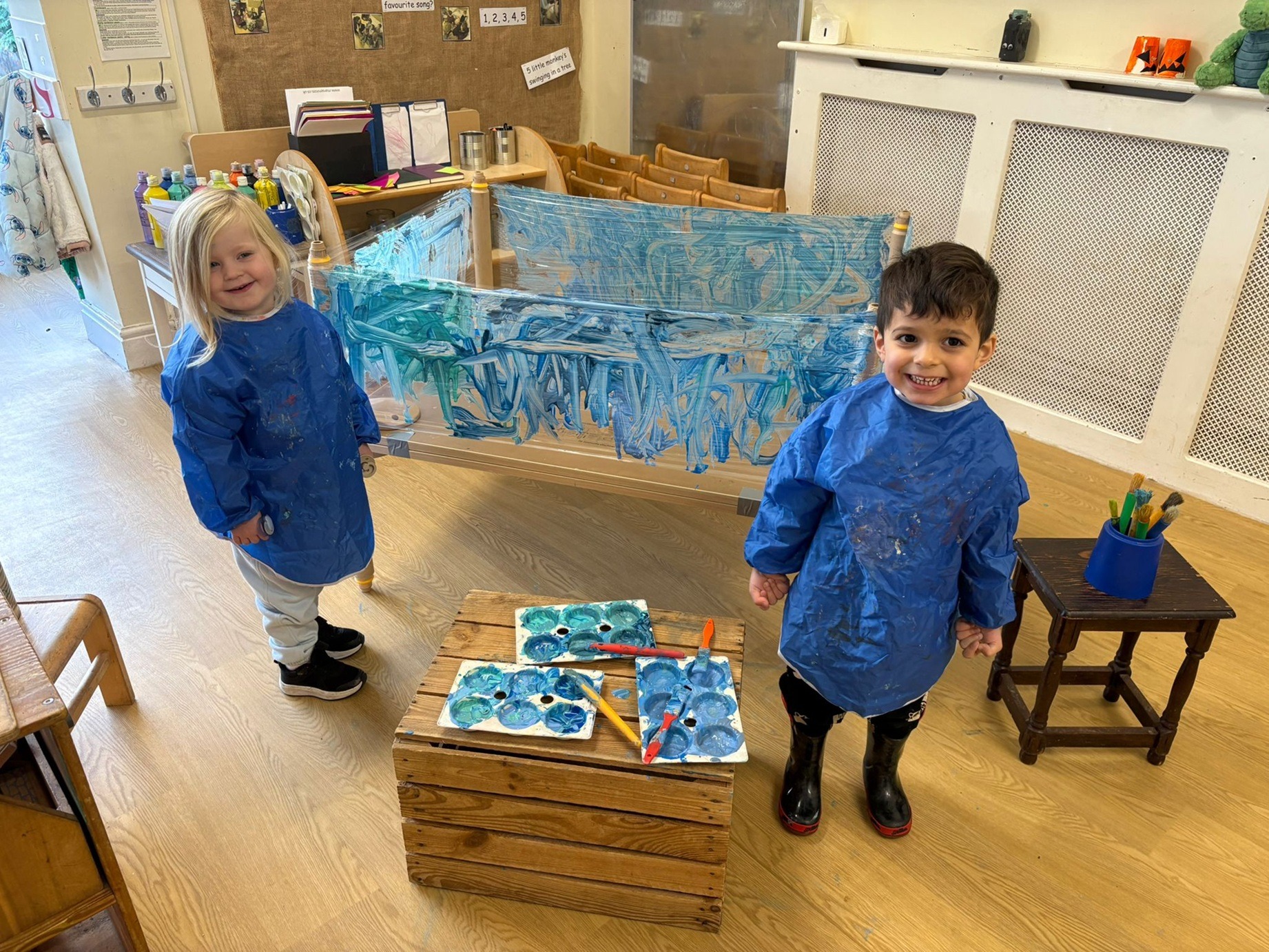 A blonde girl and brown haired boy dressed in blue next to blue art creations at Partou Children's House Day Nursery