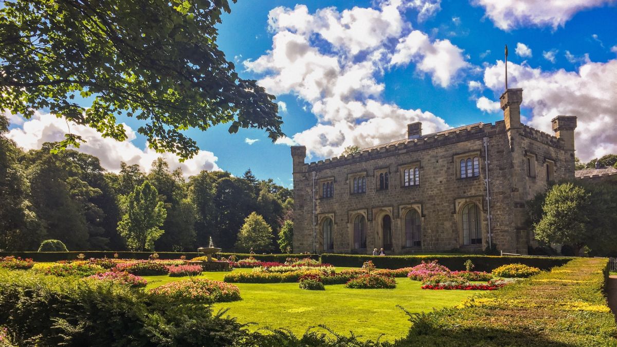 Towneley Hall in Burnley next to a large grass area with sunlight spilling over it and a beautiful big blue sky with a few white fluffy clouds