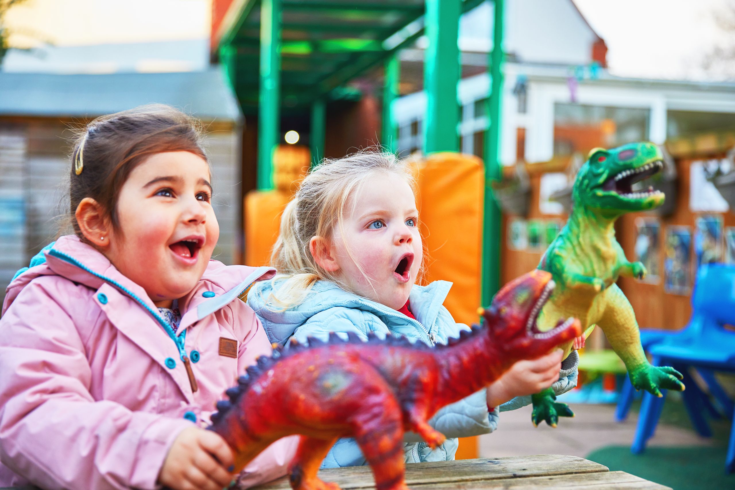 Two little girls with blonde and brown hair in brightly coloured coats playing with red and green dinosaurs outdoors at a Partou nursery and pre-school