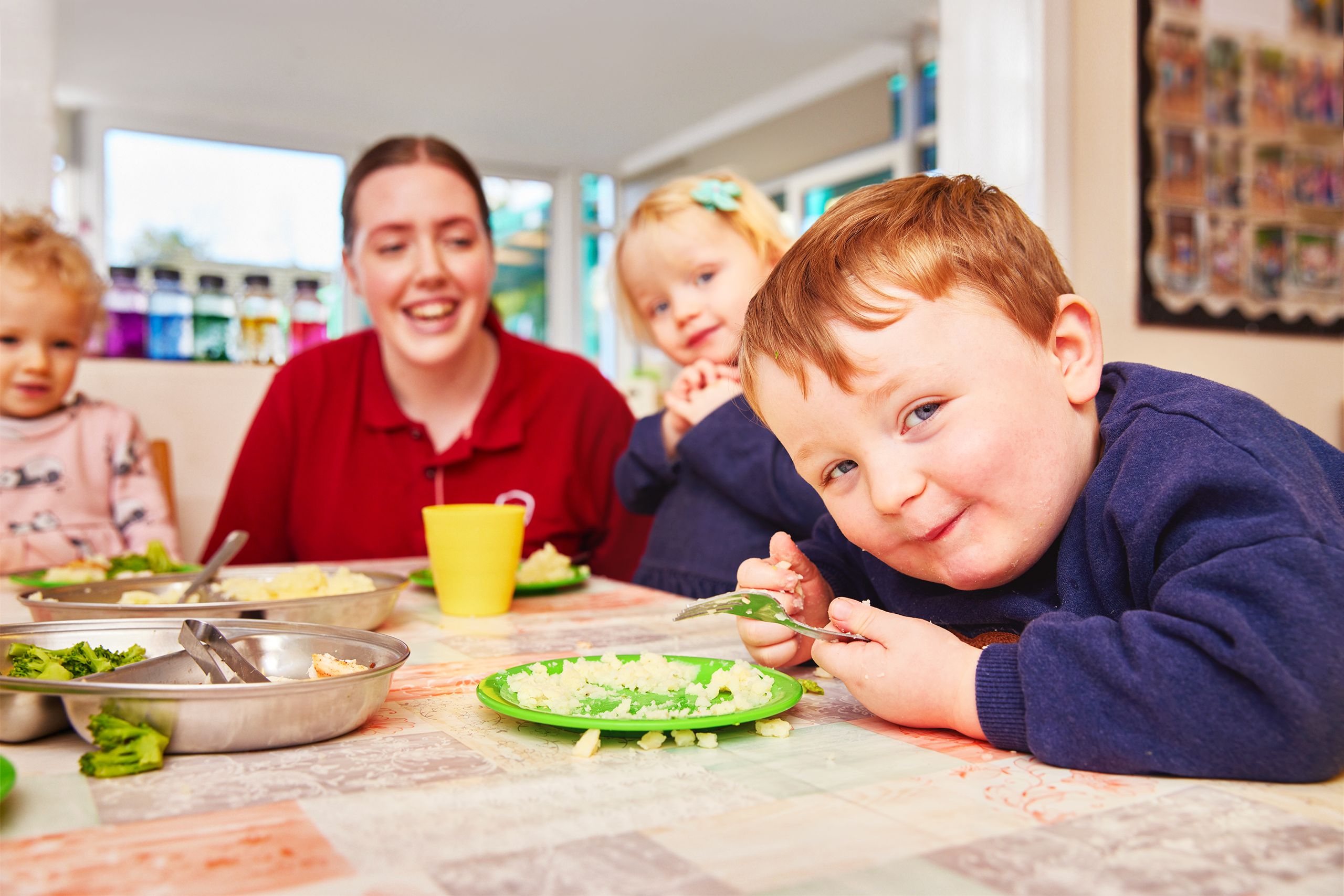 Red haired little boy in a blue jumper with a cheeky smile next to a blonde little girl with a blue flowery clip in her hair, a smiling brunette Partou team member and curly blonde haired girl wearing a pink top with pandas on. They are at the lunch table eating mashed potato and broccoli from green plates