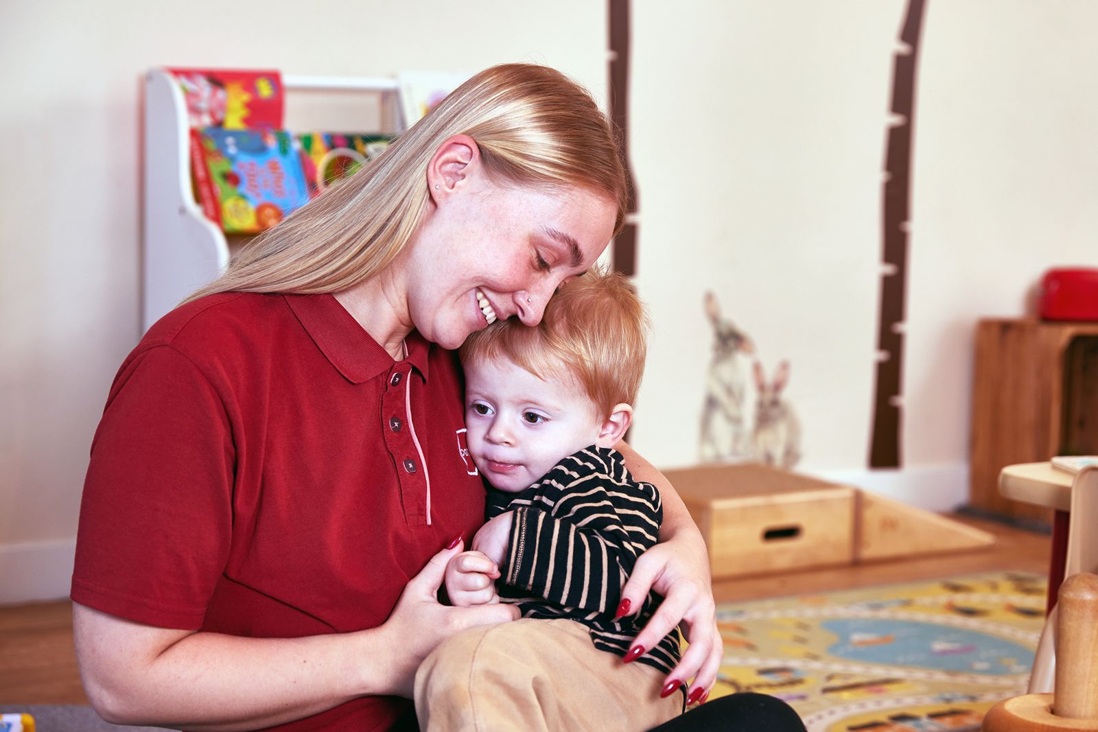 Blonde haired, female Partou nursery team member with nose and ear piercings and red nails sat on the floor hugging a little boy wearing a black and beige striped long sleeved t-shirt with beige trousers surrounded by natural resource materials and books