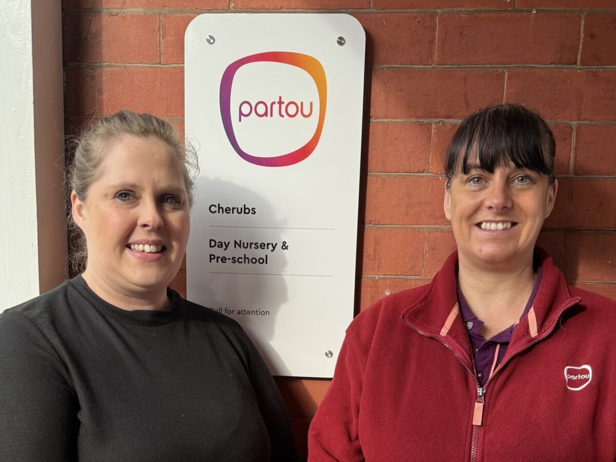 Two nursery team members pose outside a Partou day nursery, standing next to a Partou sign on a brick wall, marking a positive Ofsted inspection outcome.