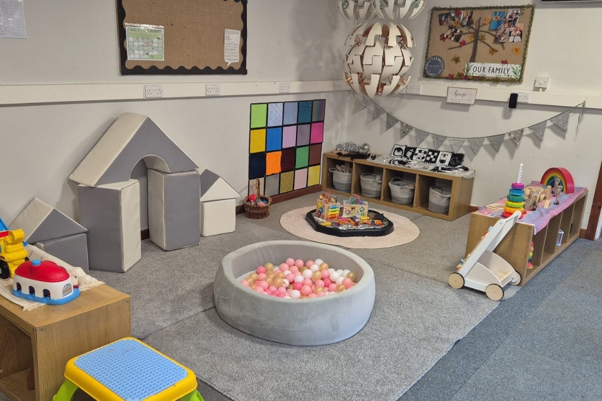 A bright, well‑organised playroom at Partou Brinscall Day Nursery & Pre‑School, featuring soft play blocks, a small round ball pit with pink, white and gold balls, a textured sensory wall panel, and neatly arranged shelves of toys and baskets, with family displays and bunting creating a welcoming, play‑based learning environment.