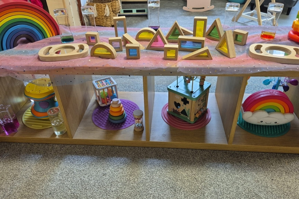 A neatly organised wooden shelf at Partou Brinscall Day Nursery & Pre‑School, displaying colourful wooden sensory blocks with transparent centres, liquid‑filled tubes and a range of early‑years toys including a drum, stacking rings, a bead‑maze cube and a rainbow toy, creating an inviting, visually stimulating space for exploratory play.