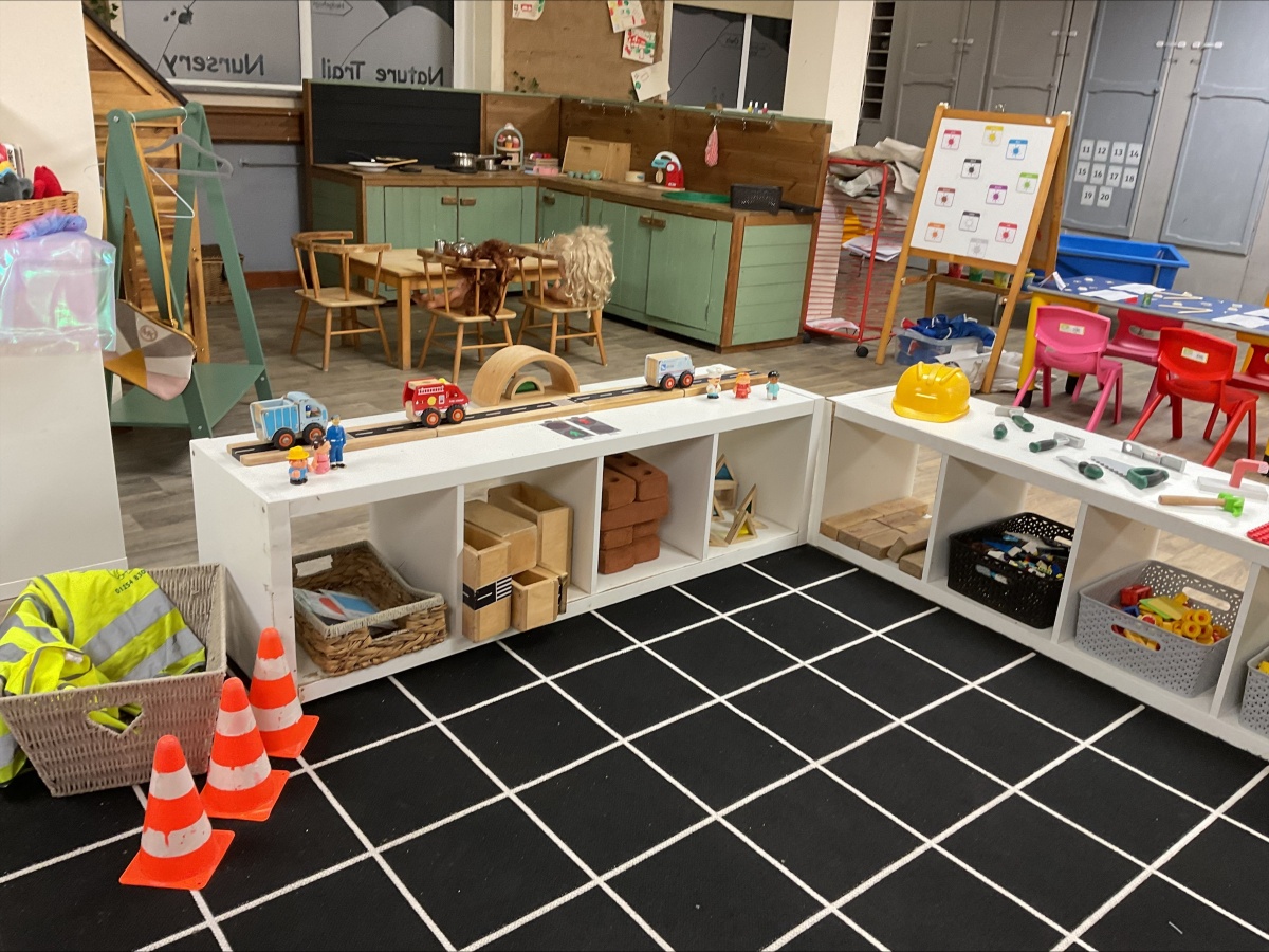 Construction and small-world learning zone at Partou Brinscall Day Nursery, featuring white modular shelving with woven baskets and loose-part materials, wooden railway track with red fire engine and vehicles, bright yellow hard hat, neon-yellow and red high-visibility jacket and traffic cones, natural wooden dining table with chairs, sage-green kitchen cabinetry with black chalkboard, black and white geometric floor tiles, visual timetable display, pink and red seating areas, and varied small-world props, encouraging imaginative play, problem-solving and early years learning through construction scenarios.