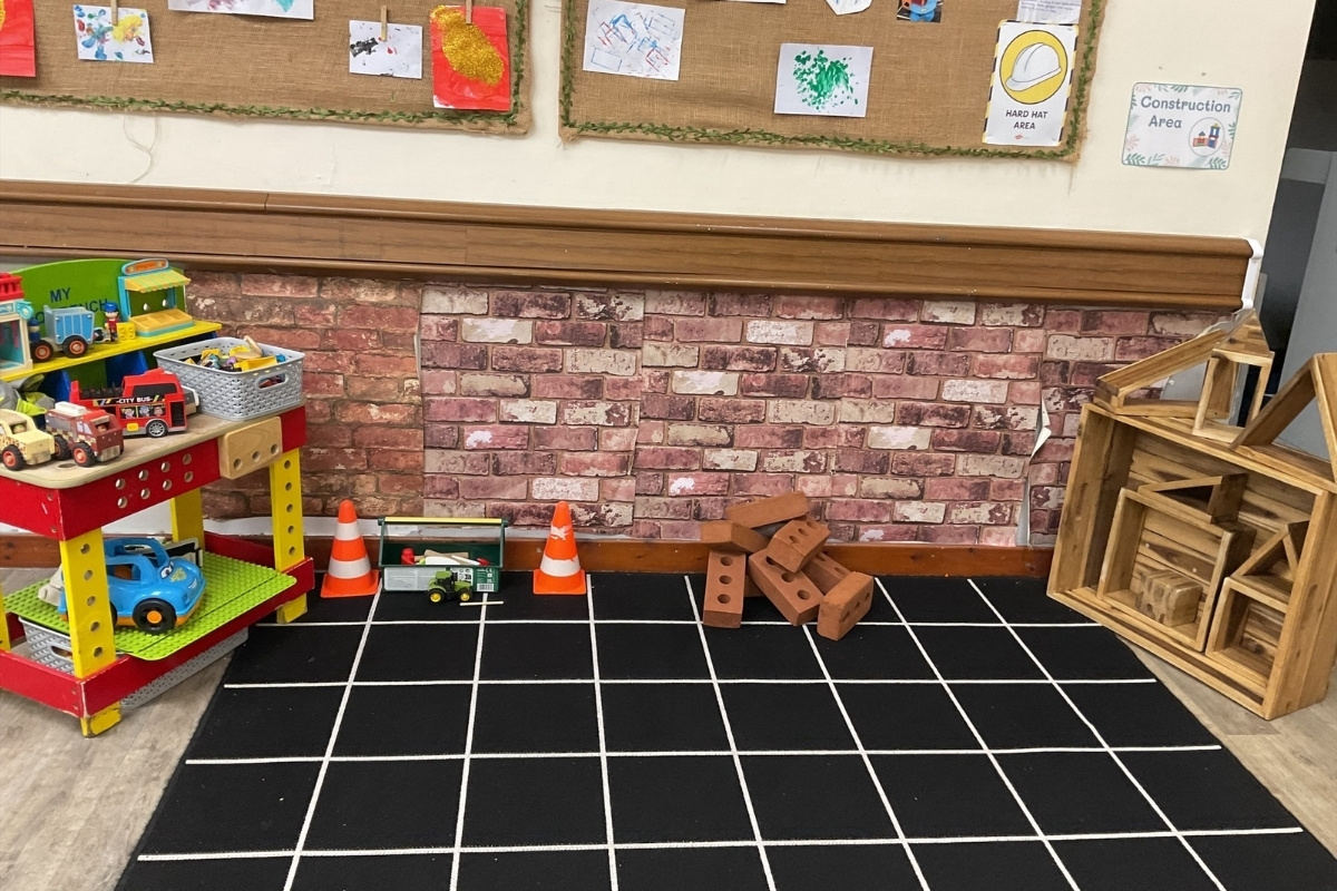 Construction area at Partou Brinscall Day Nursery, featuring realistic brick wall backdrop in terracotta and cream tones, black and white geometric play mat, red and yellow multi-level tool workbench with vehicles and accessories, natural wooden building blocks and brick-shaped loose parts, orange traffic cones, green small-world digger, natural wooden storage shelving, cork display boards with children