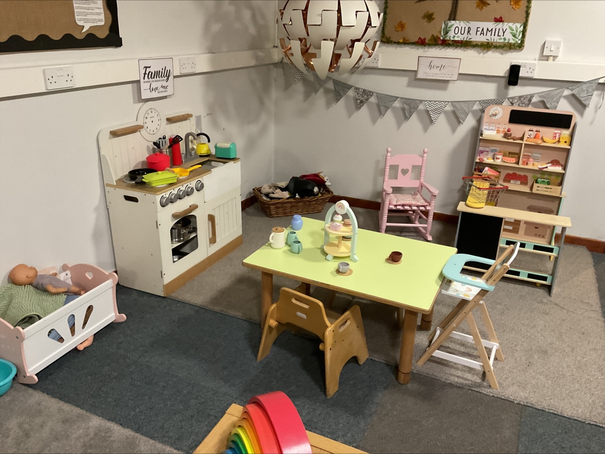 Comprehensive home corner at Partou Brinscall Day Nursery, featuring cream and natural wood play kitchen with red kettle and yellow cookware, pink wooden high chair, lime-green topped dining table with natural wooden chairs, natural wooden storage shelving with play food and accessories, cream and grey decorative bunting, "Family" and "Our Family" wall signs, woven storage basket, charcoal-grey carpet, cream walls with wooden picture rail, and varied small-world props including baby doll and pram, supporting imaginative family role-play, practical life skills and emotional development in the early years classroom.