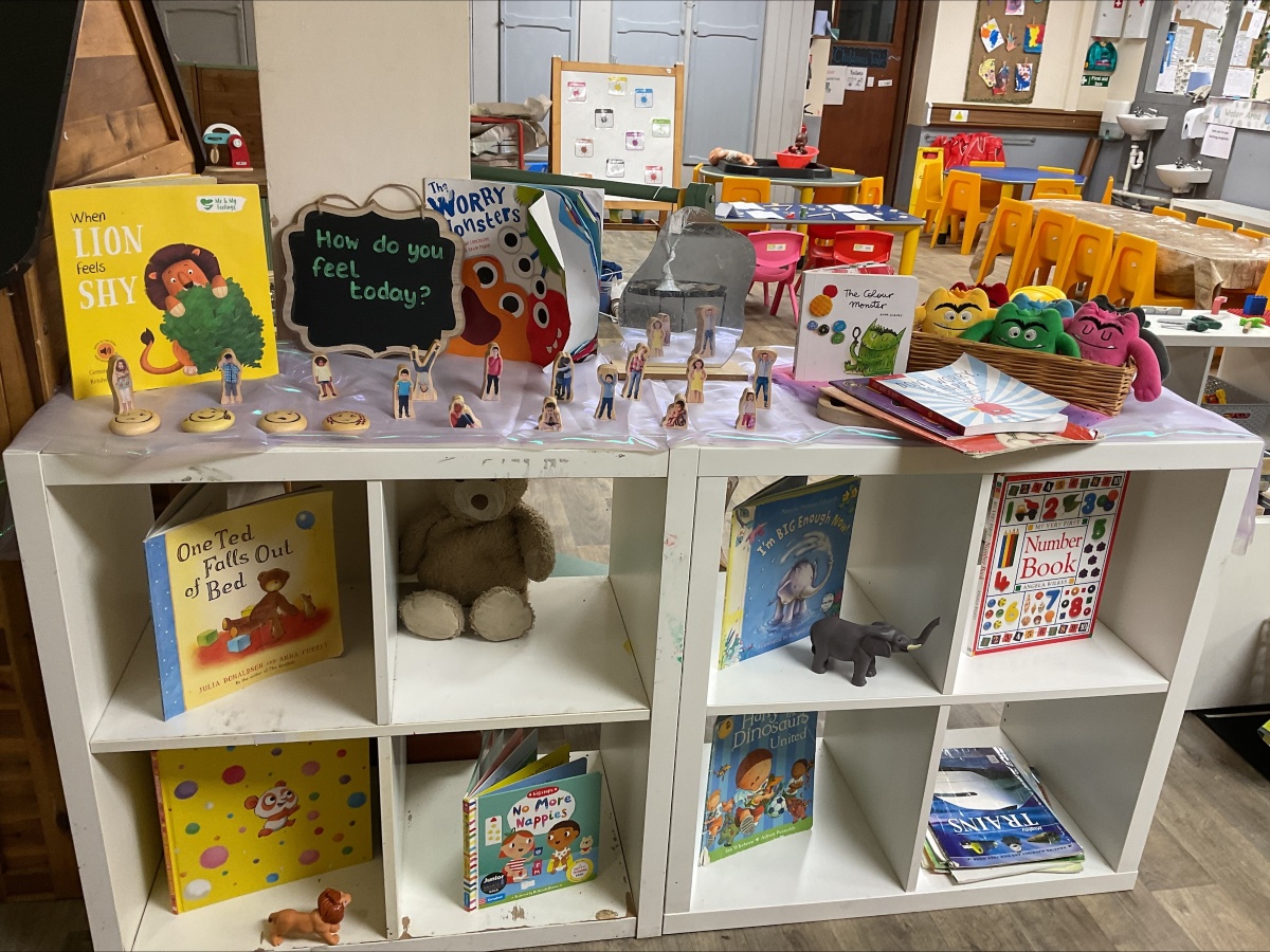 Literacy and emotional wellbeing learning zone at Partou Brinscall Day Nursery, featuring white open shelving with picture books including "When Lion Feels Shy," "One Egg Fell Out of Bed," and "The Worry Monsters," wooden character figurines on white platform, black chalkboard with "How do you feel today?" prompt, colourful puppet characters in pink, green and orange, soft grey toy elephant, children