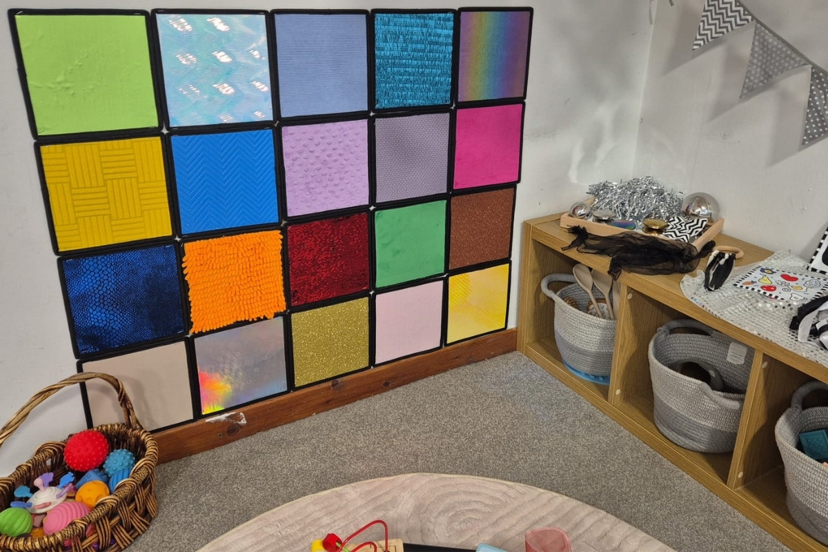 A colourful sensory area at Partou Brinscall Day Nursery & Pre‑School, featuring a wall of textured squares in bright colours, a wooden shelf with baskets of sensory materials, shiny and reflective objects displayed on top, and a wicker basket of textured balls beside a circular rug with a bead‑maze toy, creating an inviting space for tactile and visual exploration.