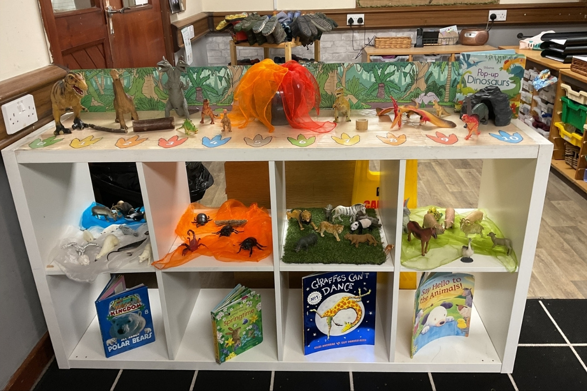 Nature and animal discovery zone at Partou Brinscall Day Nursery, featuring white open shelving with themed small-world habitats, bright orange and red silk fabric canopy with dinosaur figures and natural wooden logs, jungle-themed backdrop with green foliage, colourful paper leaf cutouts in rainbow colours, animal figurines including dinosaurs, lions, giraffes and safari creatures, thematic picture books including "Giraffes Can