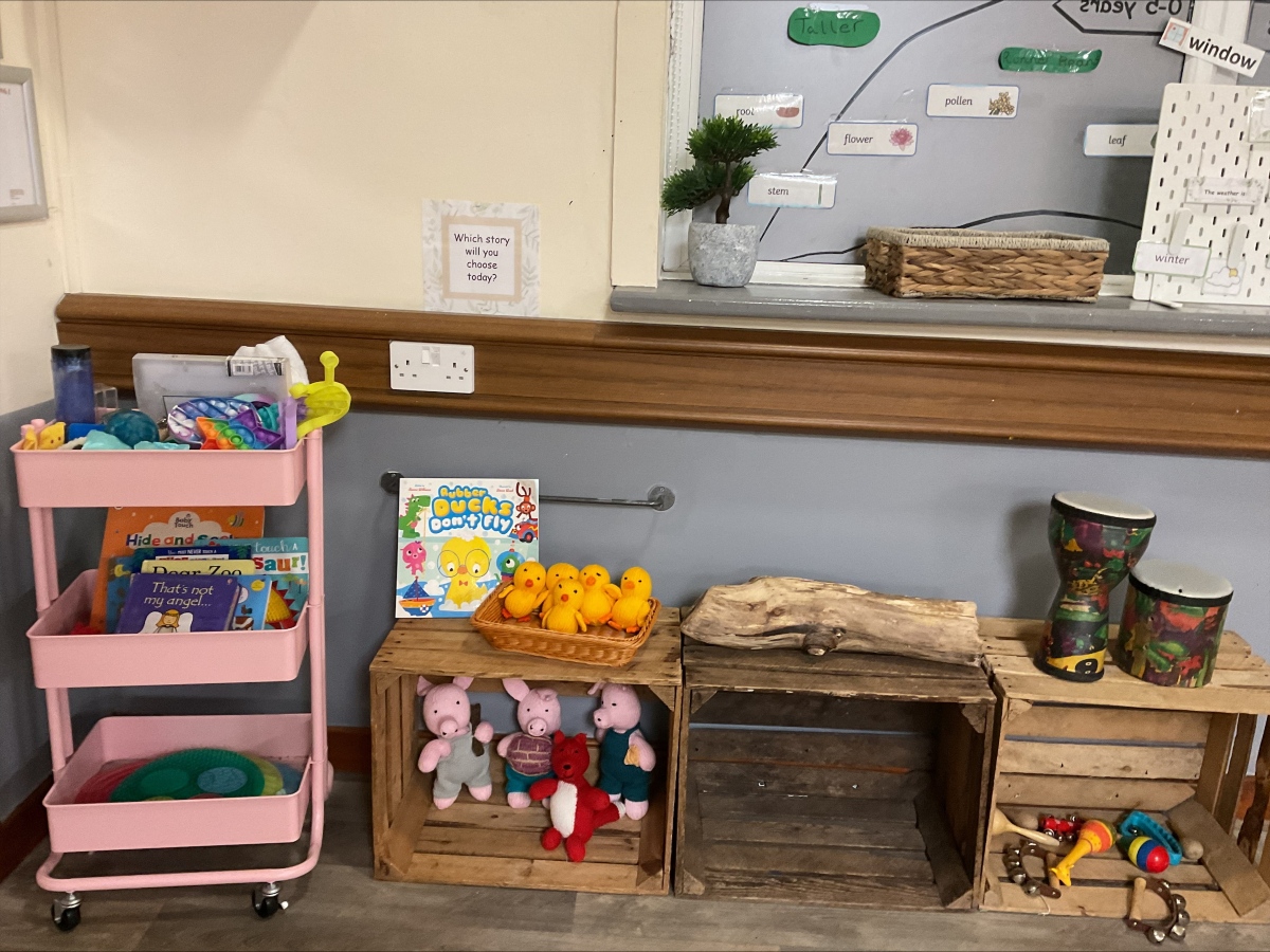 Literacy and nature learning station at Partou Brinscall Day Nursery, featuring pink rolling storage trolley with picture books including "Hide and Seek," "Dear Zoo" and "I am my Angel," grey wall display with plant-part labels (flower, stem, leaf, pollen), potted green plant with woven basket, natural wooden crates holding pink soft-toy animals, bright yellow chick figurines in woven basket, natural wooden logs, seasonal weather labels including "Winter," "window" prompt asking "Which story will you choose today?," colourful patterned storage containers, cream and grey walls with wooden picture rail, supporting early literacy, nature vocabulary and story choice in the early years classroom.