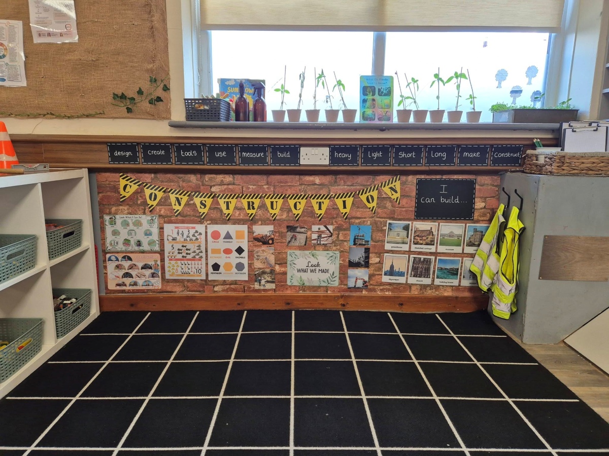 A bright construction‑themed learning area at Partou Brinscall Day Nursery & Pre‑School, featuring a brick‑patterned display wall with vocabulary cards, shape and number posters, a “I can build…” inspiration board, child‑sized high‑vis vests on hooks, and a windowsill with seedlings, books and baskets set up for imaginative building play.
