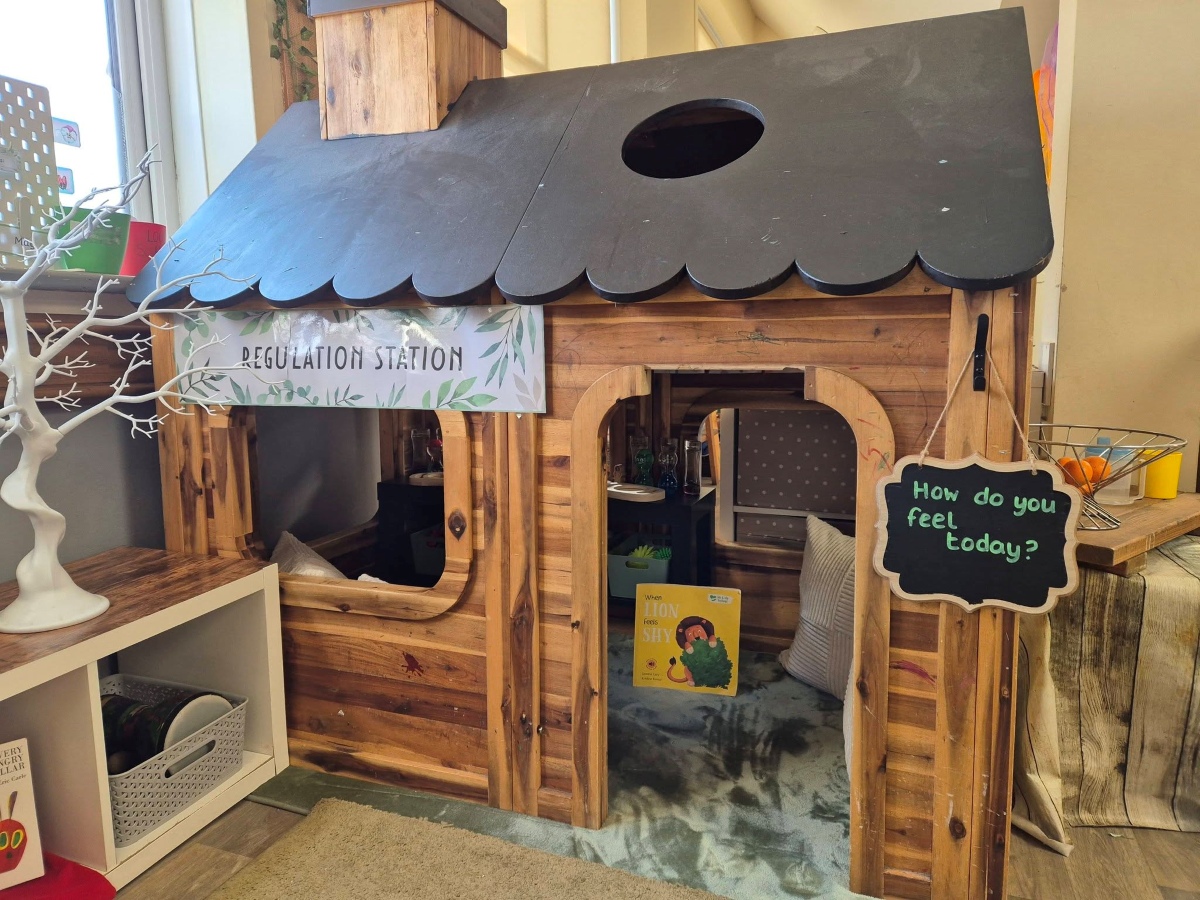 A cosy wooden “Regulation Station” playhouse at Partou Brinscall Day Nursery & Pre‑School, with soft cushions, a feelings check‑in sign, and the book The Lion Inside inside, surrounded by calming shelves and sensory items to support children’s emotional regulation.