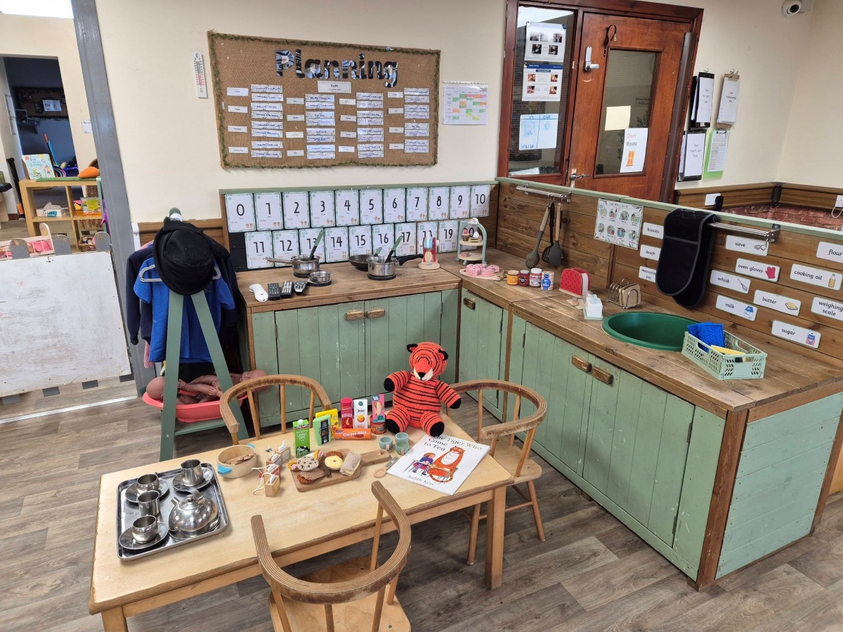 A welcoming home‑corner kitchen at Partou Brinscall Day Nursery & Pre‑School, with green wooden cabinets, play pots and utensils, labelled ingredient cards, a planning board and number line on the wall, and a small table set with play food and a soft toy tiger for imaginative cooking and role play.