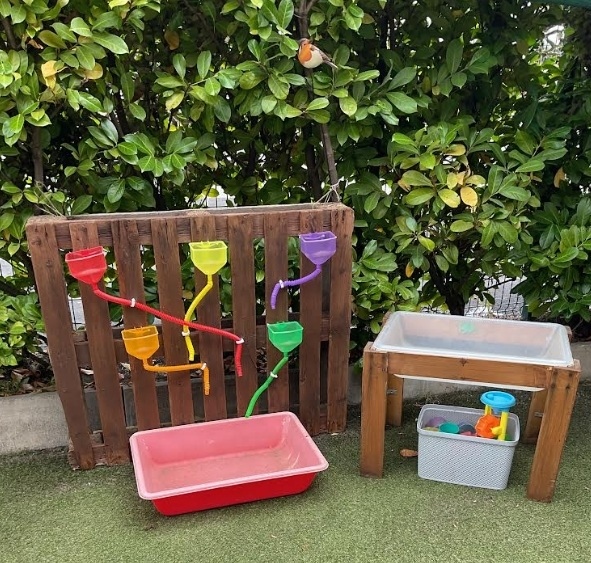 An outdoor water‑play station at Partou Chorley Day Nursery & Pre‑School, featuring a wooden pallet fitted with colourful funnels and tubes leading into a red basin, alongside a small table with a clear water tray and a basket of scoops and toys, set on artificial grass with leafy greenery behind to encourage hands‑on exploration and cause‑and‑effect learning