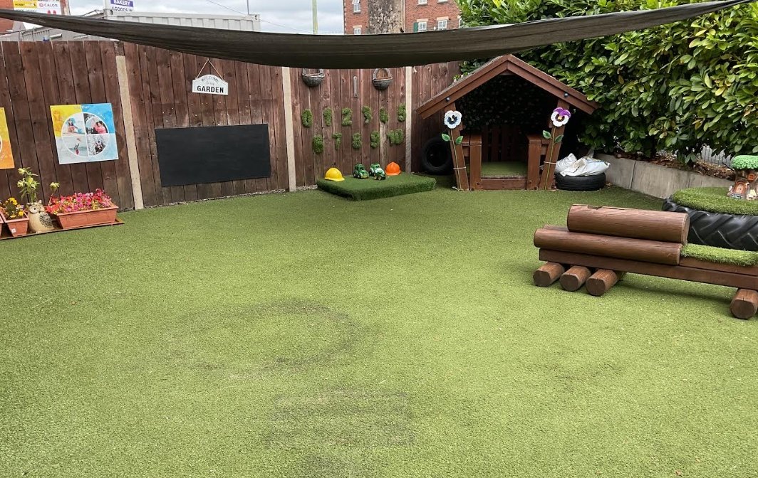 A colourful outdoor learning area at Partou Chorley Day Nursery & Pre‑School, featuring artificial grass, a wooden fence with posters and a ‘Garden’ sign, planters with flowers, a grassy construction‑play patch with helmets and vehicles, a decorated wooden playhouse, a log bench and a tyre planter, all set beneath a shade canopy to support imaginative outdoor play.