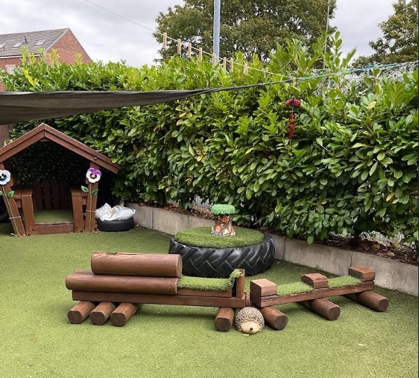 A nature‑themed outdoor play area at Partou Chorley Day Nursery & Pre‑School, featuring artificial grass, a wooden playhouse with colourful pinwheels, a large tyre filled with greenery and small animal figures, a wooden log balance beam and a hedgehog ornament, all set against a tall green hedge under a shaded canopy.