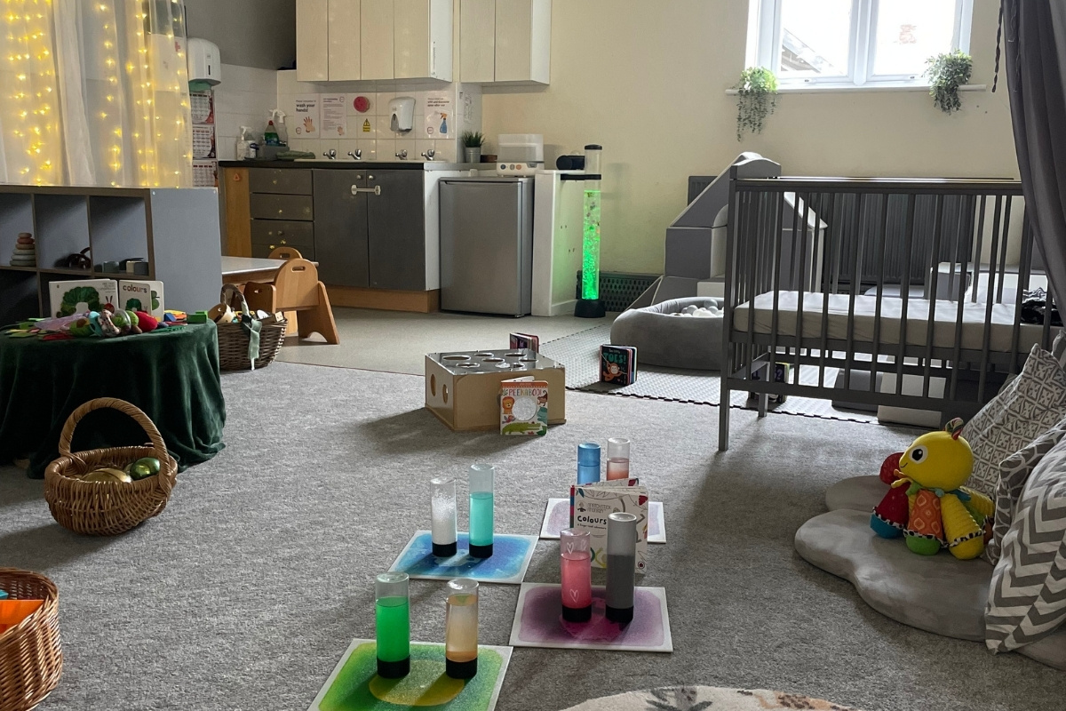 Bright, calm early years baby room at Partou Old School House in Chorley, featuring cream and soft-yellow walls with warm string lights, charcoal cot, stainless steel kitchen facilities, natural wooden high chair, green-draped activity table with woven baskets, sensory play bottles in jewel-toned greens, pinks and blues on contrasting mats, soft furnishings, hanging trailing plants, and light grey carpet, creating a nurturing space for infant development and exploration.