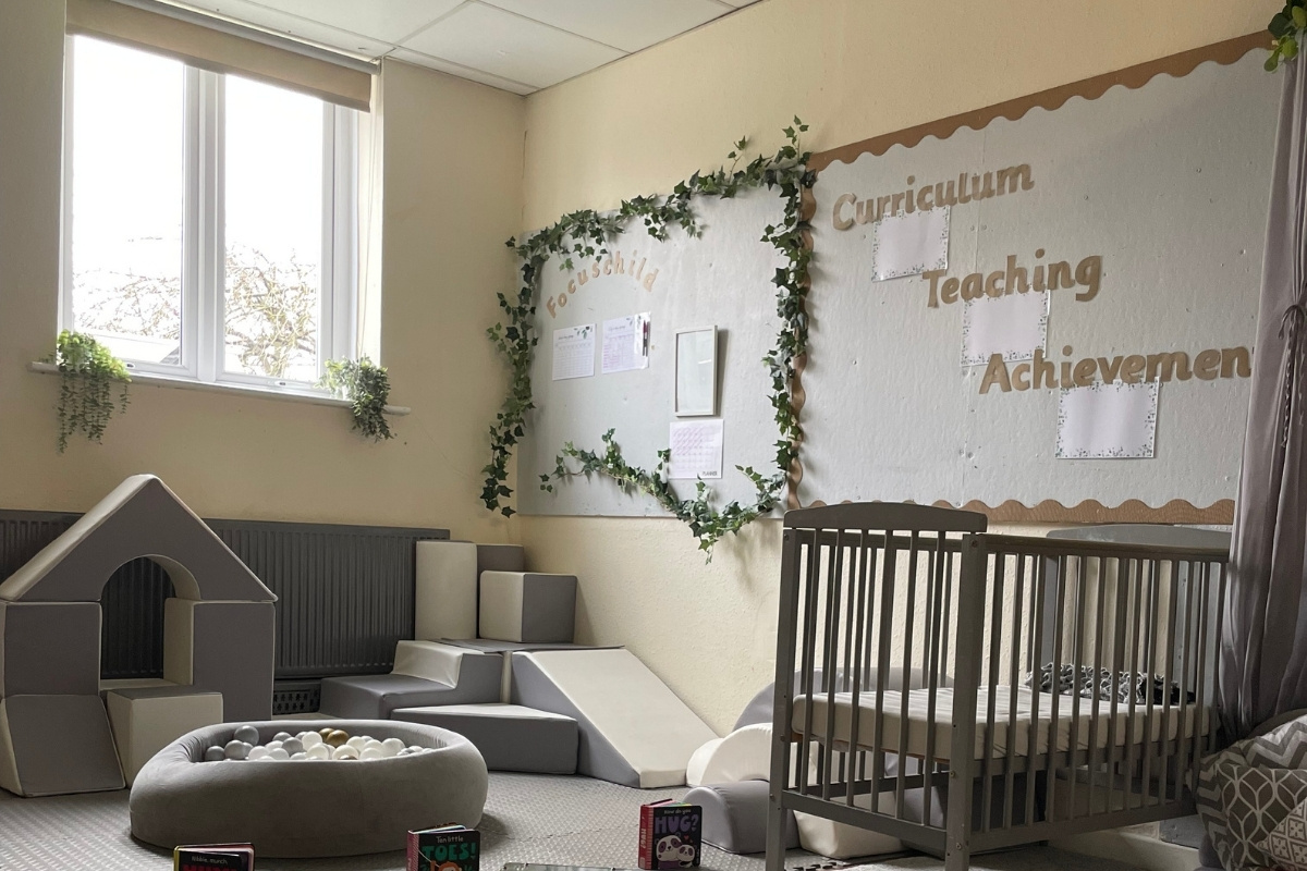 Early years classroom at Partou Old School House Chorley nursery, showcasing a calm learning environment with soft-yellow walls, charcoal half-panelling, ivy garland framing display boards, tan scalloped wall trim, text reading "Curriculum Teaching Achievement," charcoal cot, grey modular soft-play blocks, cream bean bag, grey and cream playhouse, natural light from large windows, and trailing plants on sills, supporting child development through intentional design and accessible learning spaces.