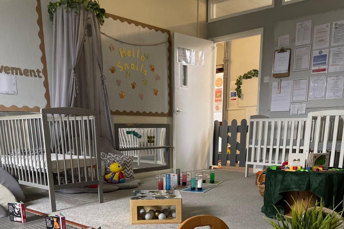 Welcoming baby room at Partou Old School House Chorley day nursery, featuring soft cream and grey walls with tan scalloped trim, "Hello Snail" handprint display with coloured handprints, white and charcoal cots, grey curtains with ivy garland, sensory play bottles in jewel-toned greens, blues, pinks and reds on white mat, wooden ball storage unit, yellow and red soft toy, charcoal gate divider, trailing plants, and curriculum documentation boards, creating a nurturing early years environment for infant care and multi-sensory learning.