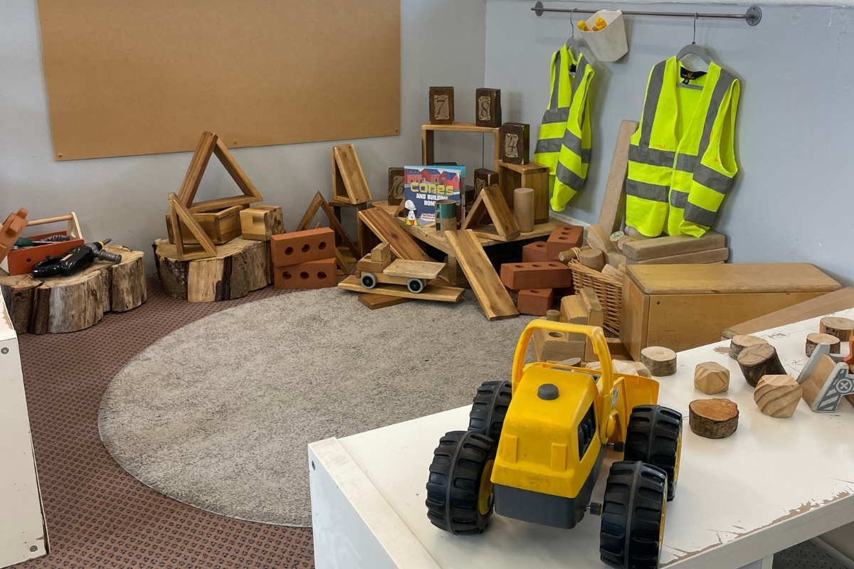 Construction and builder-themed learning space at Partou Old School House Chorley day nursery, featuring natural wooden blocks in varied shapes and sizes, wooden tree-trunk stumps, terracotta-coloured clay bricks, bright yellow digger vehicle with black tyres, natural wood circles and geometric shapes, neon-yellow and grey high-visibility jackets on wall rail, cork display board, grey carpet, burgundy patterned floor border, metal rail with storage, and loose-parts materials, encouraging spatial reasoning, imaginative play and construction skills in the early years environment.