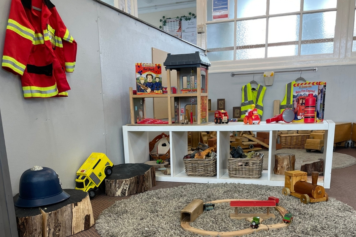 Emergency services role-play zone at Partou Old School House Chorley day nursery, featuring bright red fire service costume with neon-yellow reflective stripes, red wooden fire station building with black roof, red toy fire engines and accessories, navy-blue police helmet, yellow digger vehicle, natural wooden tree-trunk stumps, white shelving unit with woven storage baskets, large windows with natural light, grey patterned carpet, and varied small-world props, encouraging imaginative community role-play, heroic narratives and social understanding in the early years classroom.