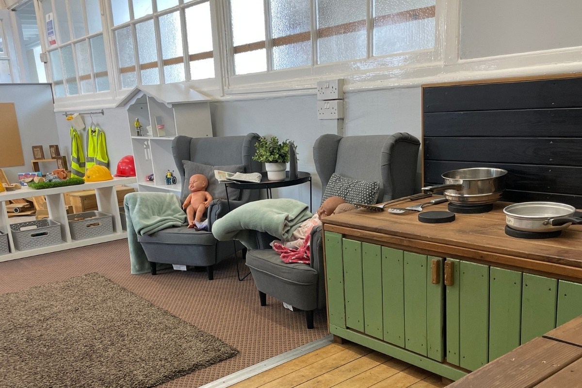 Nurturing home environment at Partou Old School House Chorley nursery, featuring grey upholstered armchairs with sage-green blankets and baby doll, natural wooden kitchen unit with green cabinetry and stainless steel cooking vessels, black wall-mounted shelving, potted trailing plant, neon-yellow high-visibility jackets on white wall shelving, red and yellow hard hats, cream and charcoal carpet, large windows with natural light, and comfortable seating areas, supporting emotional security, imaginative family role-play and early years wellbeing.