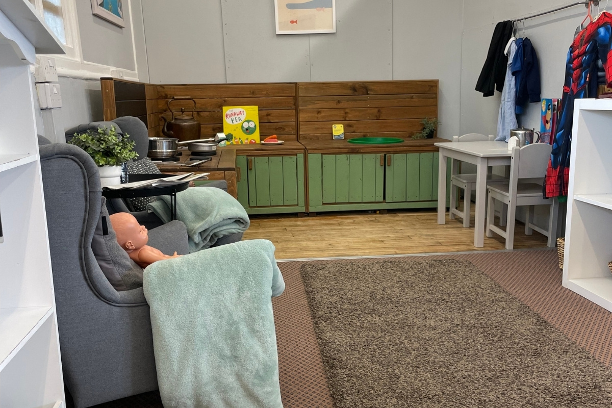 Multi-purpose home and learning environment at Partou Old School House Chorley day nursery, featuring grey upholstered armchair with sage-green blanket and baby doll, natural wooden kitchen unit with green cabinetry, stainless steel cookware and "Grumpy Frog" book, potted trailing plant, cream and green storage units with clothing rail, small wooden table and chairs, warm wooden flooring, beige and charcoal patterned carpet, large window, and varied learning materials, supporting emotional attachment, imaginative play and flexible early years provision.