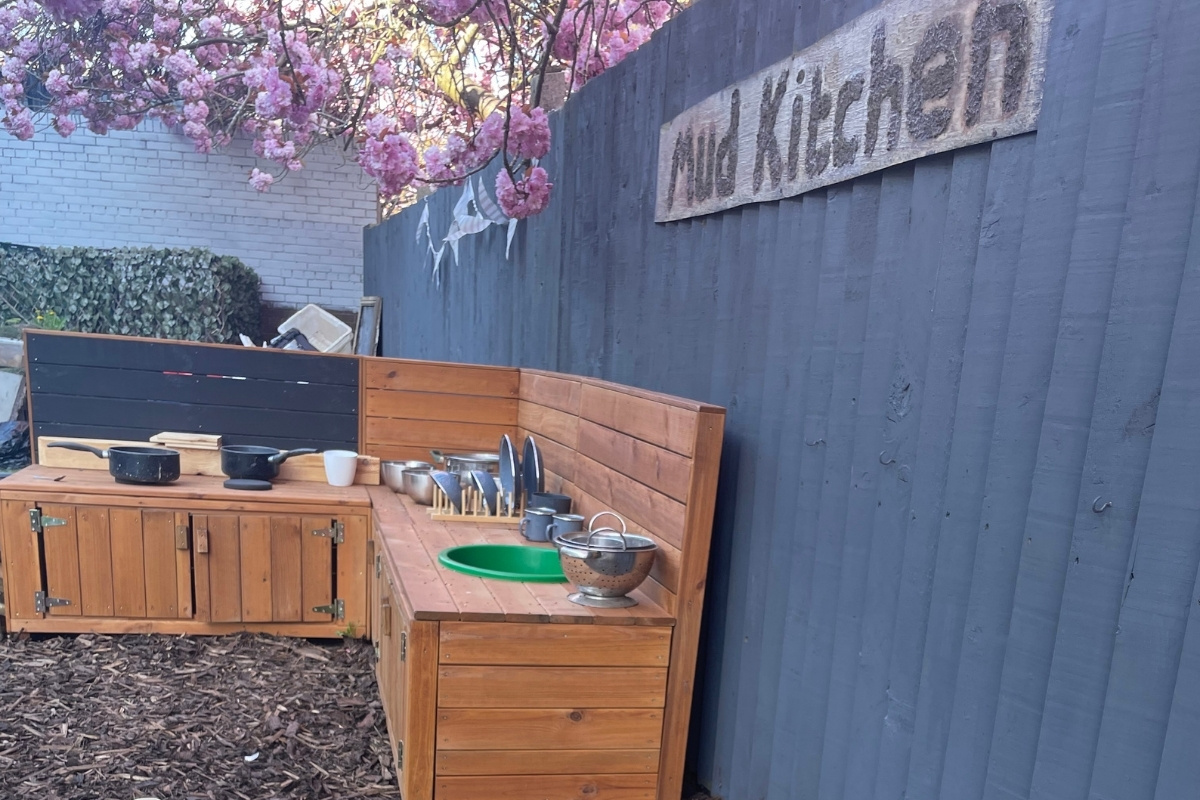 Outdoor learning kitchen at Partou Old School House Chorley nursery, featuring natural wooden countertops and storage units with metal cooking vessels, bright green plate, white crockery, stainless steel cutlery, charcoal wooden back panel, slate-blue rendered wall, vibrant pink magnolia blossom overhead, dark wood-chip ground surface, and white brick boundary, supporting practical life skills and imaginative outdoor play in the early years garden.