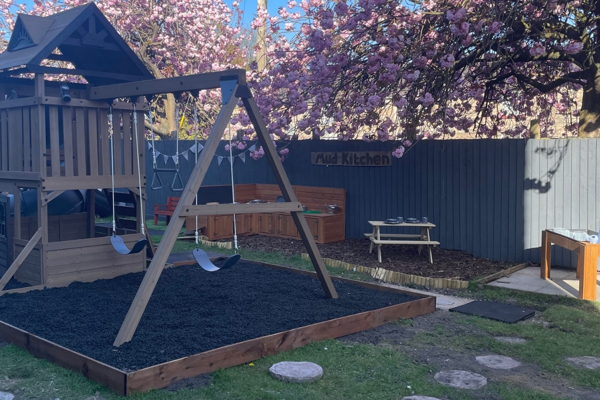 Expansive outdoor garden at Partou Old School House day nursery, featuring pale-wood play tower with blue slide, wooden swing frame with blue seats, natural wooden bench seating, charcoal-blue fencing, dark wood-chip ground surface with timber edging, vibrant pink magnolia blossom overhead, outdoor mud kitchen sign, stepping stones, and green growing areas, providing free-flow outdoor learning and physical development opportunities in the early years setting.