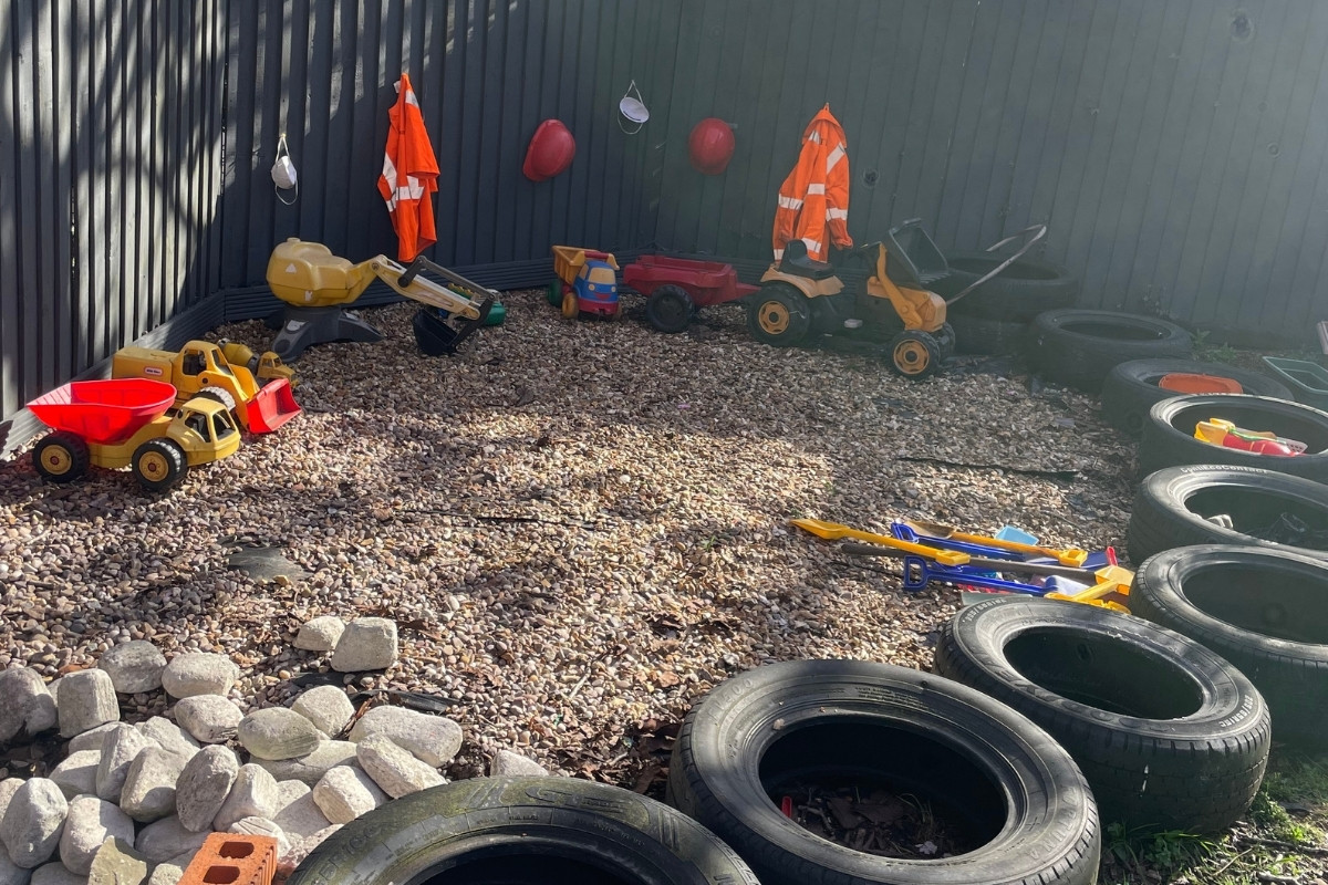 Outdoor construction play area at Partou Old School House Chorley nursery, featuring natural loose-parts learning with yellow and red digger truck, wooden wheelbarrow, colourful ride-on tractors, black tyres arranged as climbing obstacles, pale stones and gravel ground surface, bright orange and white high-visibility clothing hung on charcoal-grey fencing, red and burgundy buckets, multicoloured rescue-service toys, and reclaimed materials, encouraging gross motor skills, imaginative role-play and risk-taking in the early years outdoor environment.