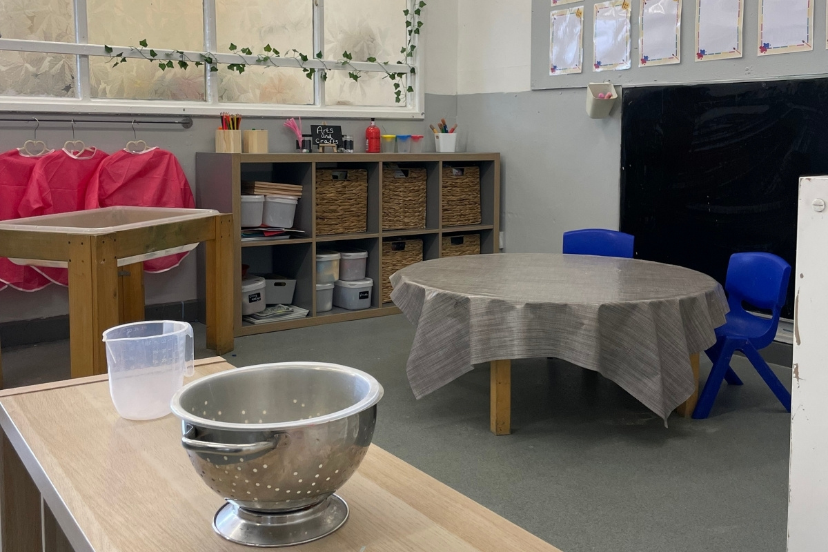 Creative and practical learning space at Partou Old School House Chorley day nursery, featuring natural wooden worktops with stainless steel colander and transparent measuring jug, woven-basket storage unit, bright magenta fabric drapes on wooden rail, bright blue plastic chairs, grey-topped table with linen cloth, "Arts and Crafts" sign, hanging ivy garland near large windows, grey-charcoal carpet, wooden shelving with art supplies and containers, and curriculum display boards, supporting sensory exploration and life skills development in the early years classroom.