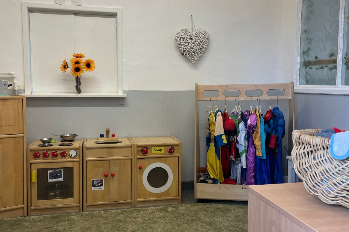 Role-play learning area at Partou Old School House Chorley nursery, featuring natural wooden play kitchen with red knobs and "Soap" label, wooden washing machine with white circular door, cream and grey half-panelled walls, dress-up rail displaying colourful jackets in yellow, red, purple, blue and pink, woven storage baskets, cream window sill with bright yellow and orange sunflowers in vase, white woven heart decoration, and soft grey carpet, supporting imaginative play, practical life skills and emotional development in the early years setting.