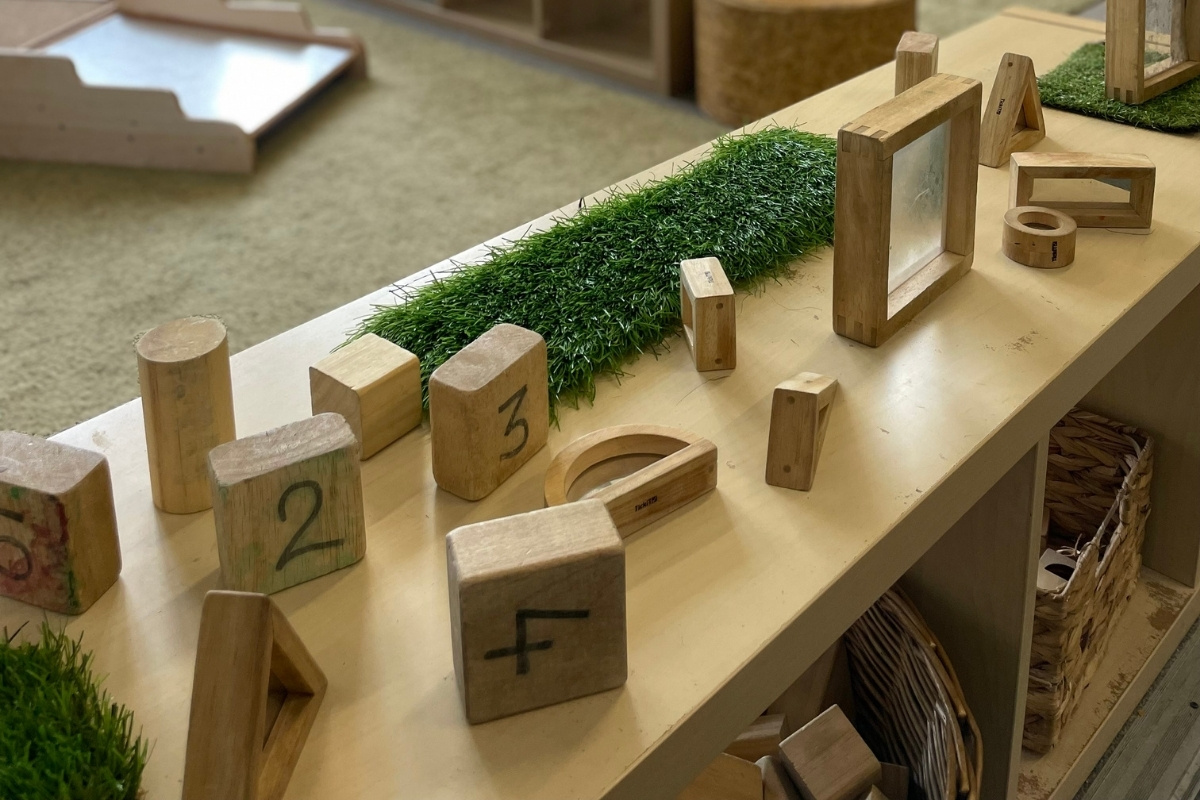 Natural loose-parts learning display at Partou Old School House Chorley day nursery, featuring pale wooden blocks numbered 1 to 4, wooden geometric shapes and frames, bright green artificial grass strip, natural wood circles and cylinders, woven storage baskets, cream shelving unit, soft grey carpet, and light-filled classroom space, encouraging mathematical exploration, fine motor development and open-ended play in the early years environment.