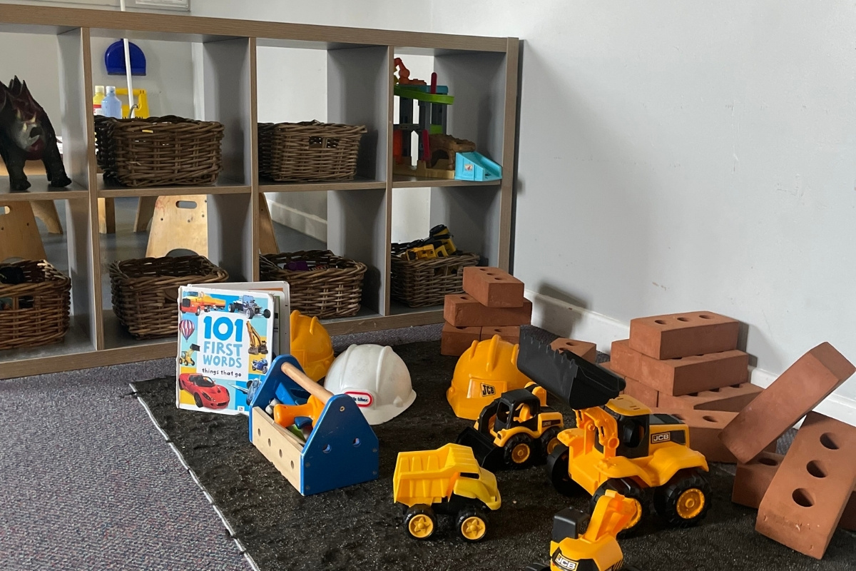 Construction and imaginative play zone at Partou Old School House Chorley nursery, featuring natural wooden shelving unit with woven storage baskets holding loose parts and toys, bright yellow JCB diggers and dumper trucks, terracotta-coloured building bricks, white hard hat and blue safety jacket, "101 First Words" book, charcoal grey carpet, cream walls, wooden workbench with blue vice, and varied small-world resources, encouraging role-play, problem-solving and vocabulary development in the early years classroom.