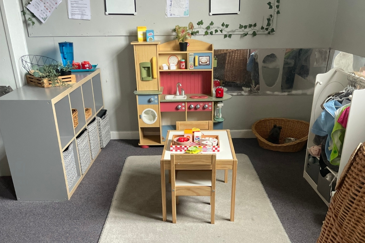 Domestic role-play kitchen at Partou Old School House Chorley day nursery, featuring natural wooden play kitchen with red cooker, blue oven, white sink, cream crockery and utensils, wooden dining table with checkered place mat, woven storage baskets, grey and charcoal-blue cabinetry, cream carpet with grey charcoal border, potted trailing plants, washing machine unit, laundry basket, curriculum documentation boards, and hanging ivy garland, supporting practical life skills, collaborative play and early years learning through real-world kitchen and home scenarios