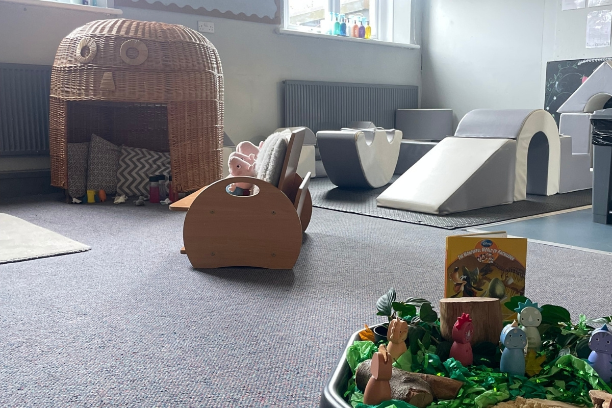 Sensory and gross motor learning area at Partou Old School House Chorley nursery, featuring woven wicker pod den with chevron-patterned cushions, chocolate-brown soft-play tunnel, cream foam climbing blocks, grey padded soft-play shapes, charcoal radiator, natural light from large windows, nature-themed small-world tray with green moss, wooden animal figurines and story book, grey carpet, and varied tactile materials, supporting physical development, imaginative exploration and calming spaces in the early years environment.