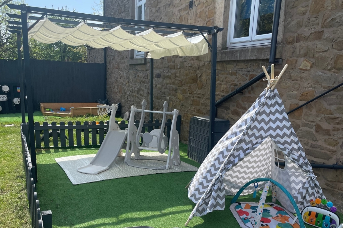 Secure outdoor garden at Partou Old School House Chorley day nursery, featuring cream canopy shade structure with charcoal-blue pergola frame, grey and white chevron-patterned play teepee with wooden poles, pale-grey climbing frame with slide, natural green lawn, raised wooden sandpit, charcoal-blue fencing, warm sandstone building wall, and sensory play mat, providing safe outdoor learning, physical activity and imaginative play in the early years garden.