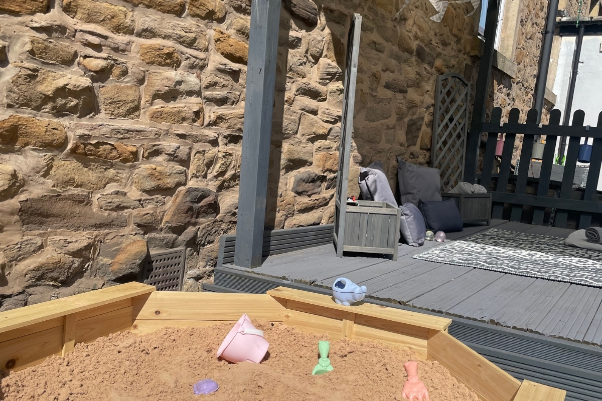 Outdoor sandpit and quiet garden at Partou Old School House Chorley nursery, featuring natural wooden sand table with pale sand and coloured play objects in pink and mint-green, warm honey-coloured sandstone building wall, charcoal-grey decking, black picket fencing, grey storage chest with cushions, patterned play mat, and sheltered patio area, providing sensory sand play, quiet exploration and cosy outdoor spaces for early years learning and relaxation.