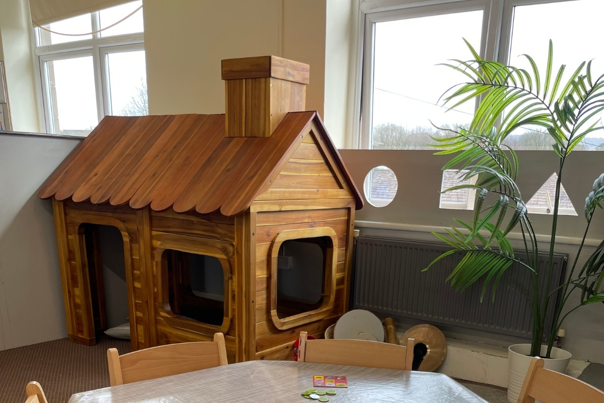 Imaginative play cottage at Partou Old School House Chorley nursery, featuring natural wooden playhouse with pitched roof, shingle detailing and multiple window openings, light and mid-tone wood construction, potted bright-green trailing plant with striped fronds, natural wooden dining table and chairs, cream crockery and play food, charcoal radiator, soft-grey walls with large windows providing natural light and outdoor views, and warm wood flooring, encouraging role-play, spatial exploration and cosy learning in the early years environment.