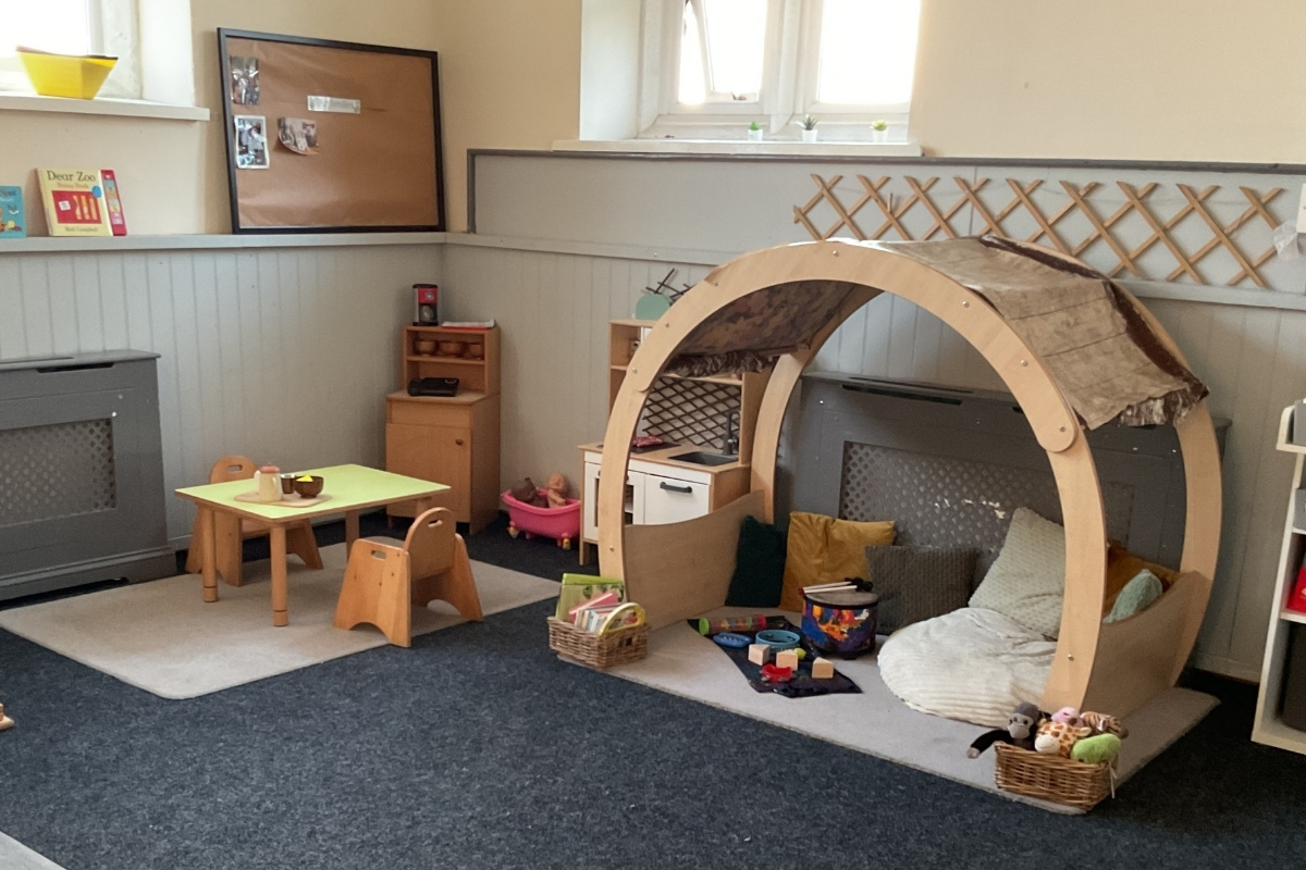 Bright, welcoming early years classroom at Partou Parbold Day Nursery in Wigan, featuring a natural wooden pod den with cream upholstered seating, a lime-green-topped activity table with wooden stools, charcoal-grey carpet, cream half-panelled walls, cork display board, storage baskets, and picture books, designed to support independent play and cosy learning spaces for children in Wigan