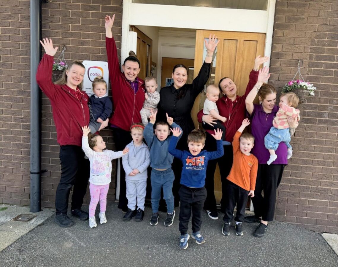 Team members and children from Partou Upton Day Nursery & Pre‑school in Birkenhead pose outside the nursery entrance with raised hands during an Ofsted celebration.