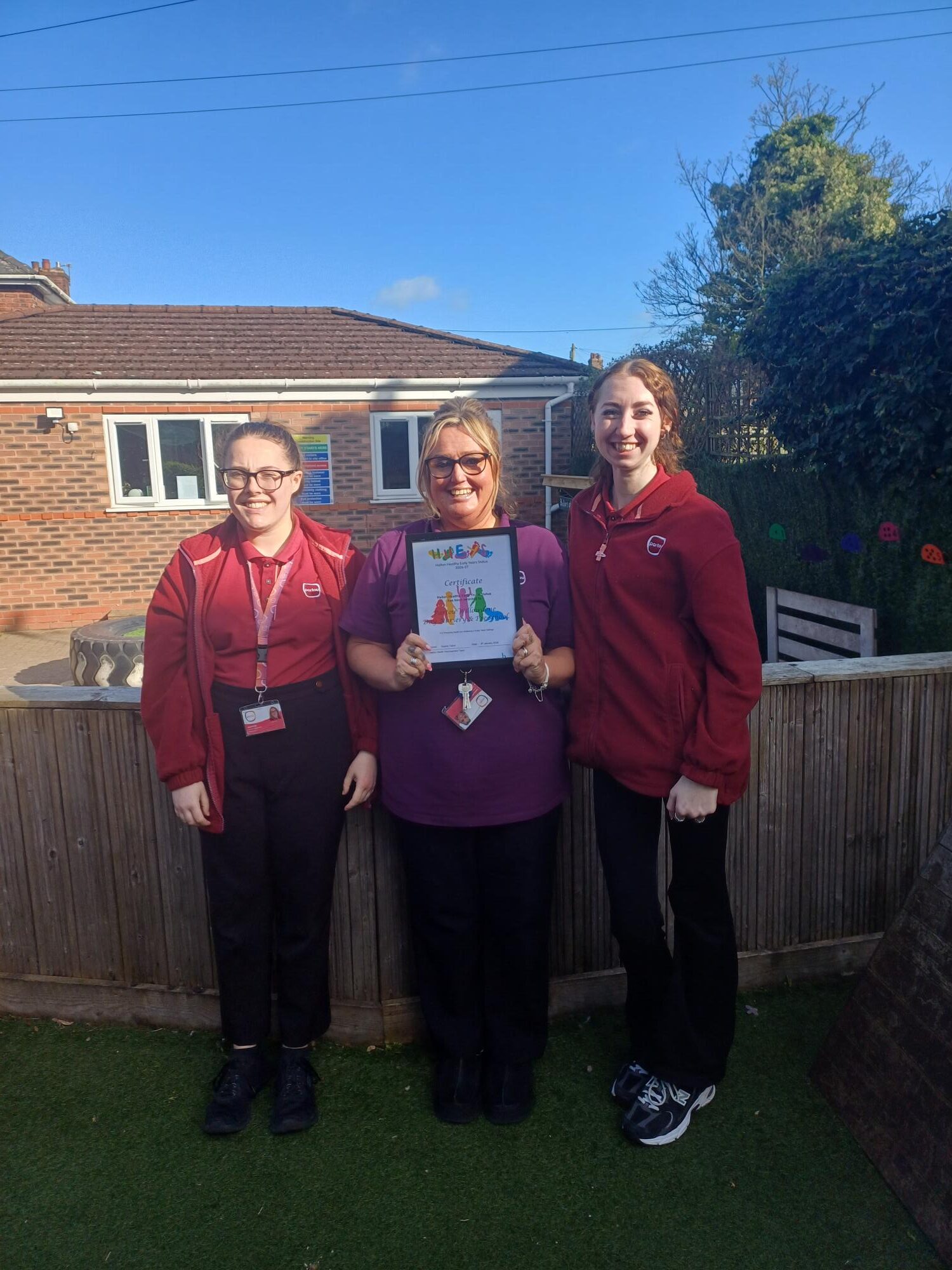 Three female nursery practioners from Partou Willowdene in Widnes standing outside next to a fence holding their Halton Healthy Early Years certificate
